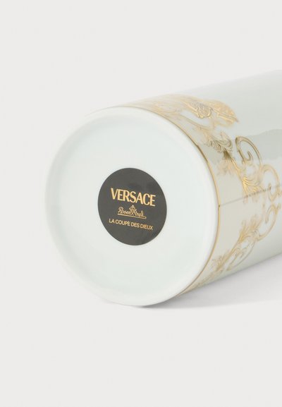 Vase en céramique blanche avec des motifs dorés ornés et une étiquette circulaire noire à la base portant l'inscription Versace Rosenthal La Coupe Des Dieux.