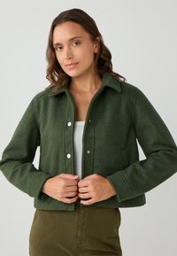 Donna con capelli castano mossi indossa una giacca verde scuro con bottoni sopra un top bianco e pantaloni verde oliva, mentre aggiusta la parte anteriore della giacca.