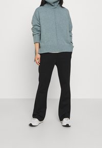 Persona vestida con un suéter de cuello alto azul claro, pantalones negros y zapatillas blancas, de pie frente a un fondo blanco liso.