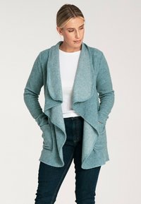 Jasnoniebieski, strukturalny kardigan z otwartym przodem i zwisającym kołnierzem. Posiada długie rękawy oraz dwie przednie kieszenie. W połączeniu z białą koszulą i ciemnymi dżinsami.