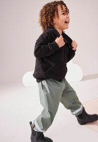 Hanorac negru cu buzunar frontal, pantaloni impermeabili verde deschis cu dungi reflectorizante și cizme de cauciuc negre. Copil zâmbind și mergând.