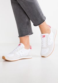 Baskets blanches avec des accents roses et des détails en daim gris, dotées d'une semelle extérieure en gomme texturée, d'un design à lacets et d'un logo "N" bien visible.