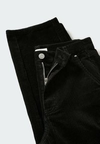 Pantalon en velours côtelé noir plié avec fermeture éclair partiellement ouverte, bouton visible et stries verticales texturées sur le tissu.