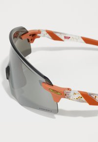 Ochelari de soare sport cu lentile gri curbate, accente din cauciuc portocalii și o ramă transparentă modelată cu stropi portocalii și galbeni.