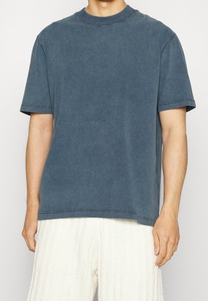 Homme portant un t-shirt bleu uni délavé à manches courtes et un pantalon blanc texturé, debout devant un fond neutre.