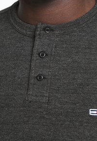 Dunkelgraues Henley-Shirt mit rundem Halsausschnitt, drei schwarzen Knöpfen und einem kleinen Logo mit horizontalen Streifen in Rot, Weiß und Blau.