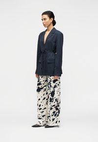 Μπλε ναυτικό σακάκι με μακριά μανίκια και ζώνη στη μέση, σε συνδυασμό με παντελόνες φαρδιάς γραμμής με ασπρόμαυρο σχέδιο πιτσιλιών. Κομψά μυτερά παπούτσια.