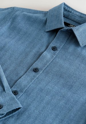 Chemise à motif chevron bleu avec un col boutonné, arborant des boutons noirs et un tissu texturé.