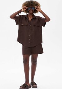 Bruine linnen shirt met korte mouwen met twee borstzakken, gecombineerd met bijpassende korte broek en chunky sandalen. Heeft een comfortabele pasvorm en knopen.