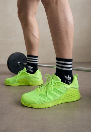 Zapatillas deportivas de color verde neón con parte superior de malla, suelas gruesas y un talón texturizado. Usadas con calcetas con rayas en blanco y negro. Barra de pesas en el fondo.