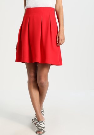 Woman wearing a knee-length red pleated skirt, white sleeveless top, and white high-heeled sandals with black stripes and bow details.