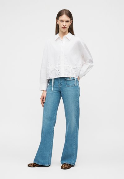 Femme portant une chemise blanche à manches longues avec des détails en œillets, un jean à jambes larges bleu clair, et des chaussures à imprimé léopard, debout avec la main sur la hanche.