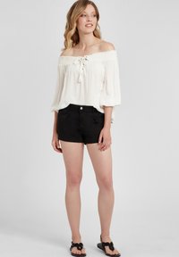 Weiße schulterfreie Bluse aus strukturiertem Stoff mit Schnürung vorne, schwarze enge Shorts und schwarze Flip-Flop-Sandalen.