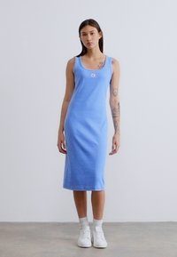 Μπλε ριμπ φόρεμα με φαρδιά τιράντα, εφαρμοστή γραμμή, μήκος μέχρι το γόνατο, με μικρό λογότυπο. Το μοντέλο φοράει λευκά αθλητικά παπούτσια και κάλτσες μέχρι τον αστράγαλο.