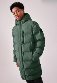 Zelena oversized puffer jakna s visokim ovratnikom, ziperom na prsnom džepu i izraženim šavovima, koja ima glatku, mat teksturu.