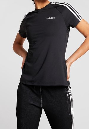 Personne portant une tenue de sport Adidas noire avec des bandes blanches sur les manches et le pantalon, debout les mains sur les hanches sur un fond gris.