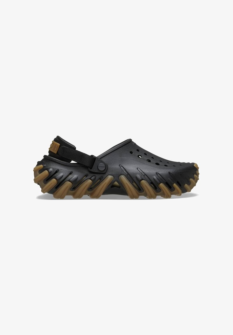 Schwarze Clogs mit einer geriffelten, braunen Sohle. Sie verfügen über Belüftungsöffnungen und einen verstellbaren Rückengurt. Hergestellt aus strapazierfähigem, strukturiertem Material.