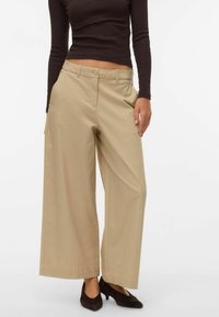 Bēša krāsas platām kājām bikses no kokvilnas, ar pogu aizdari, priekšējām kabatām un smalkām šuvju detaļām. Valkātas kopā ar melniem augstpapēžu apaviem.