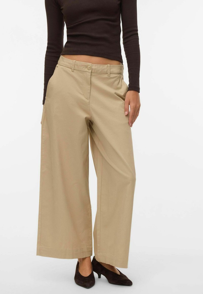 Bēša krāsas platām kājām bikses no kokvilnas, ar pogu aizdari, priekšējām kabatām un smalkām šuvju detaļām. Valkātas kopā ar melniem augstpapēžu apaviem.