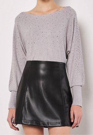 Femme portant un pull gris à manches longues avec de petits clous et une jupe en cuir noire taille haute, montrée du cou jusqu'à mi-cuisse.