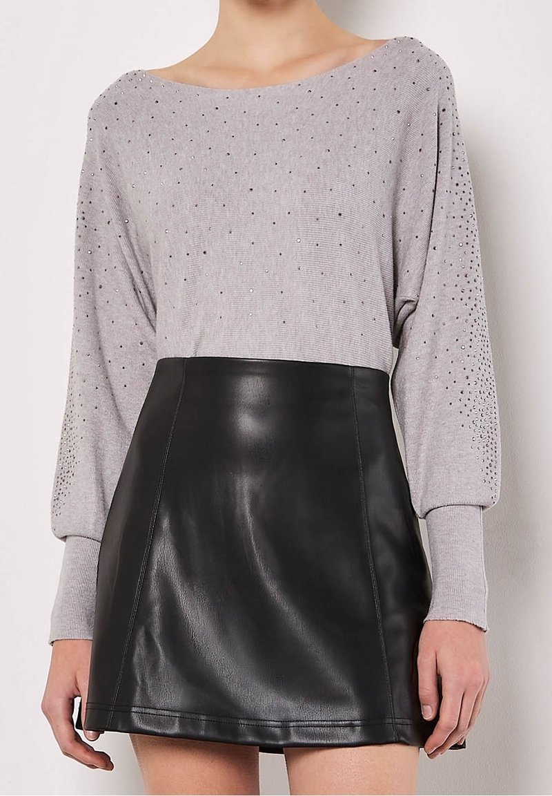 Femme portant un pull gris à manches longues avec de petits clous et une jupe en cuir noire taille haute, montrée du cou jusqu'à mi-cuisse.