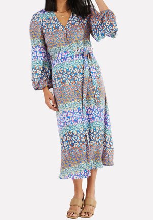 Vrouw draagt een midi-jurk met lange mouwen en bloemenprint, een strikceintuur en beige open teen sandalen, staand tegen een effen achtergrond.