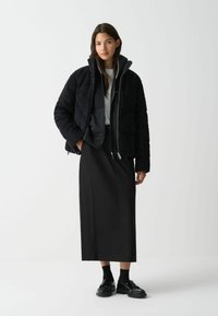Zwarte pufferjack met ribbelstructuur, voorrits en hoge kraag, gedragen over een grijze sweatshirt en gecombineerd met een zwarte rok en schoenen.