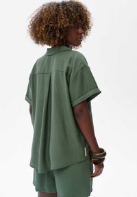 Vrouw met krullend haar staat zijwaarts en draagt een losse groene top met korte mouwen en bijpassende shorts, accessorized met armbanden om haar pols.