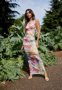 Rochie maxi florală dintr-un material transparent, cu culori vibrante și detalii din pliu. Complementată cu sandale cu toc înalt galbene.
