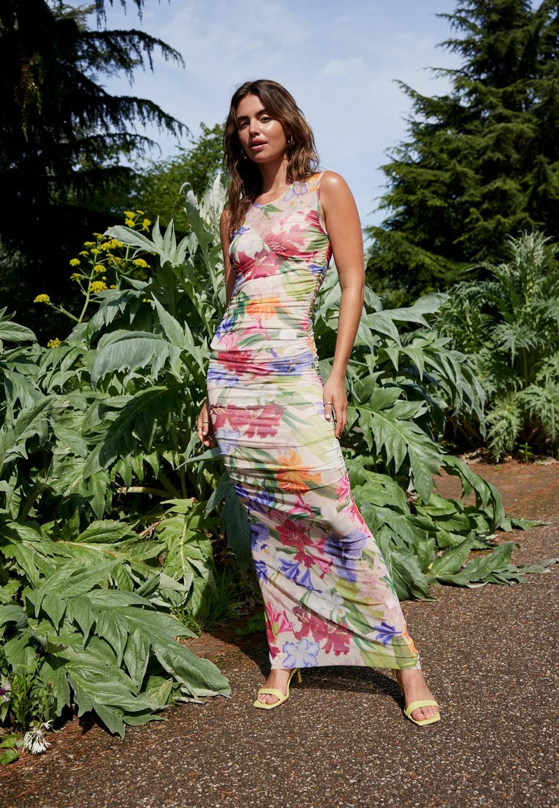 Rochie maxi florală dintr-un material transparent, cu culori vibrante și detalii din pliu. Complementată cu sandale cu toc înalt galbene.