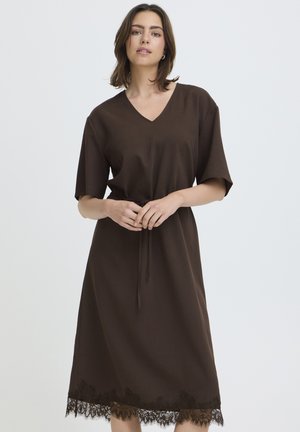 Femme aux cheveux bruns mi-longs portant une robe midi marron foncé à col en V, manches courtes et bordure en dentelle à l'ourlet, debout les mains jointes.