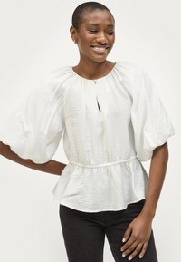 Blusa blanca de mangas abullonadas con escote redondo, detalle de ojal, cintura elástica y tejido texturado; combinada con jeans oscuros.