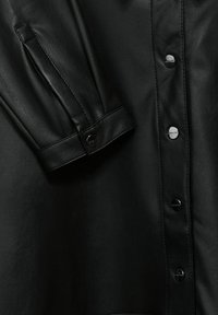 Svart läderskjorta med texturerad yta, med metalldetaljer, plymerade ärmar och en strukturerad krage. Närbild betonar detaljerna.