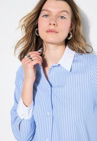 Camisa de botones de rayas finas azul claro con cuello blanco. Presenta mangas largas con puños abotonados. Textura de tela suave.