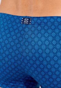 Maillots de bain à motifs bleus avec un design en losange. Présente une étiquette intitulée "VÊTEMENTS DE PLAGE" au niveau de la taille. Texture du tissu lisse.