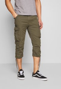 Olivgrüne Cargo-Hosen mit mehreren Taschen, verkürzter Länge, verstellbaren Bündchen und einer glatten Textur, kombiniert mit schwarzen Sneakers.