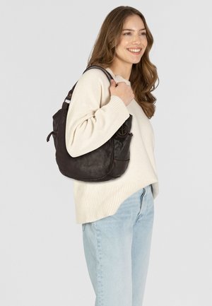 Donkerbruine leren schoudertas met een slungelige vorm, enkele strap en een externe pocket. Draagt bij een crèmekleurige trui en lichte jeans.