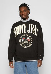 Felpa nera con audace scritta crema "TOMMY JEANS", logo di New York City, colletto con zip e polsini a costine in contrasto; indossata sopra jeans blu.