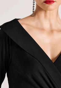 Zwarte jurk met een brede kraag en gedrapeerd ontwerp. Gladde stoftextuur. Heeft een diepe V-hals en een opvallende geometrische halslijn.