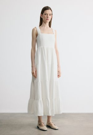 Kobieta stojąca w białej, bezrękawnej, warstwowej sukience maxi z kwadratowym dekoltem i beżowymi płaskimi butami na prostym tle.