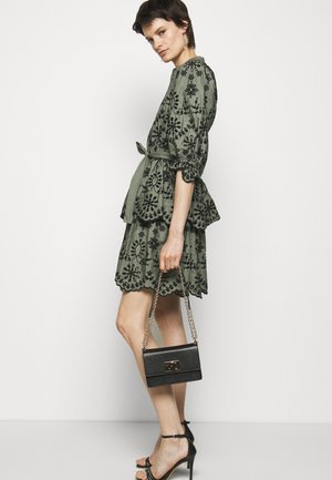 Abito verde oliva con ricami floreali neri, silhouette a peplo e maniche corte. Borsa nera a tracolla con catena e chiusura.