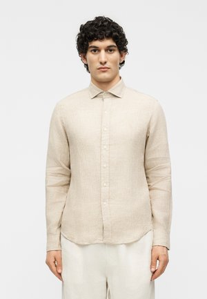 Homme aux cheveux bouclés portant une chemise beige à manches longues boutonnée et un pantalon clair, debout devant un fond blanc uni.