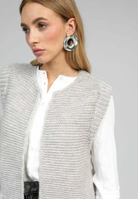 Gilets en tricot gris clair avec un motif texturé et une ouverture sur le devant. Porté sur une chemise blanche à boutons, complété par de grandes boucles d'oreilles circulaires en argent.