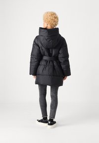 Giacca a vento nera con colletto alto, vita con cintura e design trapuntato. Presenta un cappuccio e lunghezza fino a metà coscia. Indossata con leggings grigi e scarpe da ginnastica.