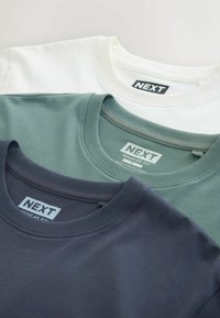 Három póló egymásra helyezve: fehér, zöld és sötétkék. Mindegyik kerek nyakkivágással rendelkezik, és egy "NEXT" feliratú címkét visel, rajta az "EGYENES VÁGÁS" megjelöléssel.