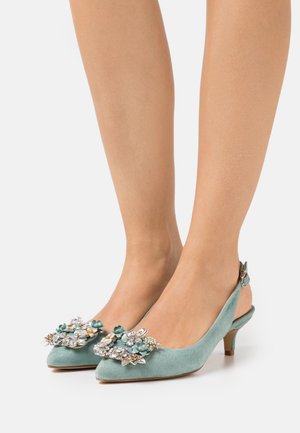 Des escarpins kitten à talons slingback en daim bleu clair avec des embellissements floraux ornés de bijoux sur les bouts pointus, portés pieds nus sur un fond blanc.