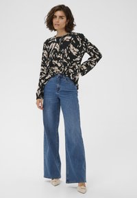 Blouse noire à motif abstrait beige, détail à boutons, manches longues, associée à un jean bleu taille haute à jambes larges et des chaussures de couleur claire.