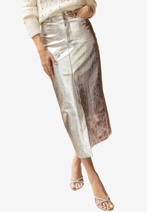 Kobieta ubrana w metaliczną srebrną spódnicę midi, biały dzianinowy sweter i srebrne sandały na wysokim obcasie z paskami, stojąca z jedną ręką w kieszeni.