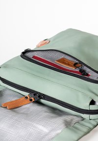 Zaino in tessuto verde con diverse cerniere, dettagli in pelle e interno grigio a quadretti. Presenta una tasca con zip rossa e un'etichetta in pelle beige.