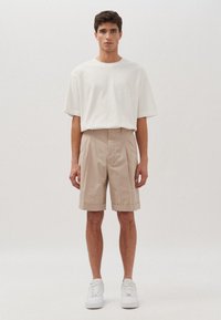 Camicia bianca a maniche corte, pantaloni corti beige plissettati e sneakers bianche indossati da un modello maschile in piedi contro uno sfondo neutro.